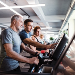 A group of seniors in gym with a young trainer doing cardio work out, exercising on stationary bicycle.